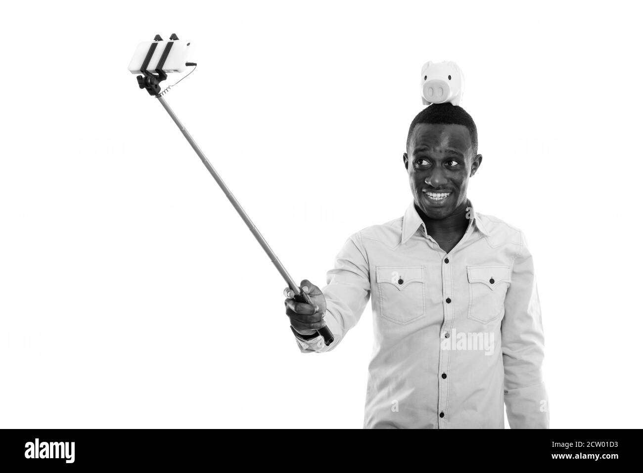 Felice giovane africano con banca piggy in cima testa prendendo selfie con il telefono cellulare su stick selfie Foto Stock