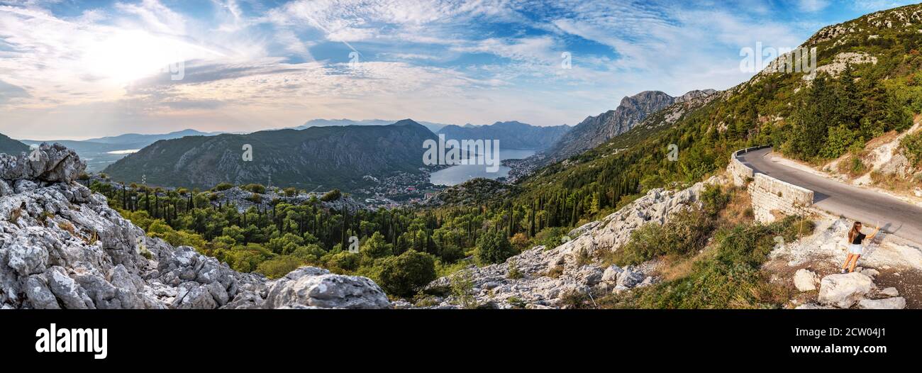 Incantevole panorama del Montenegro, città di Cattaro e baia dalla strada a serpentina di montagna. Belle montagne del Montenegro sopra il cielo con le nuvole al tramonto. Bella giovane donna che fa un selfie. Foto Stock