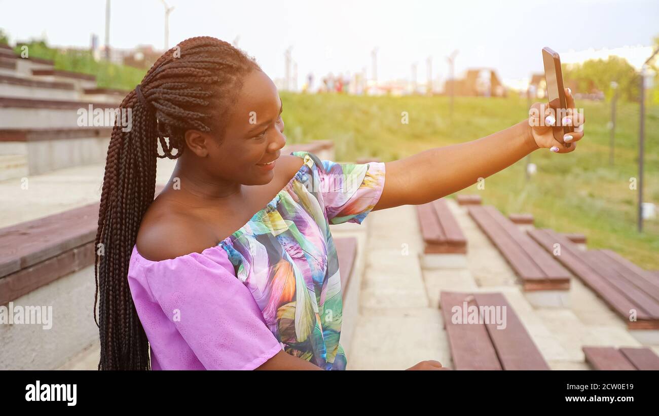 Attraente giovane afroamericana con coda lunga in colore elegante la blusa fa selfie con lo smartphone seduto sulla panca in verde parco estivo Foto Stock