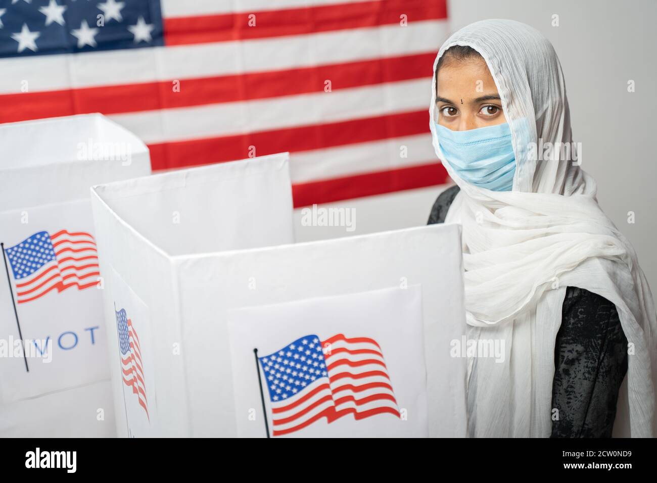 Ragazza con Hijab o copertura testa e maschera indossata alla cabina di polling guardando la fotocamera con la bandiera degli Stati Uniti come sfondo - concetto di elezione degli Stati Uniti. Foto Stock