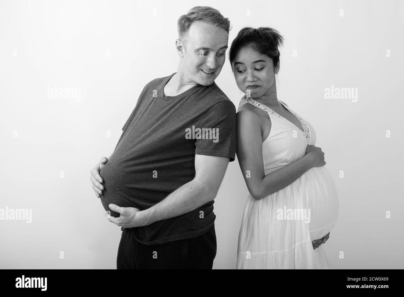 Felice giovane coppia sposata multietnica sorridente guardando lo stomaco l'uno sull'altro su sfondo bianco Foto Stock