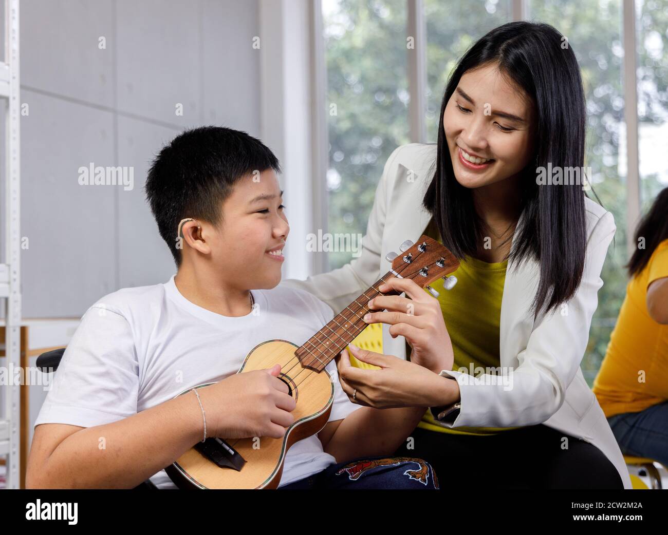 Insegnante asiatico insegnano ukulele al ragazzo disabile su sedia a rotelle. Concetto di apprendimento dei bambini disabili a scuola. Foto Stock
