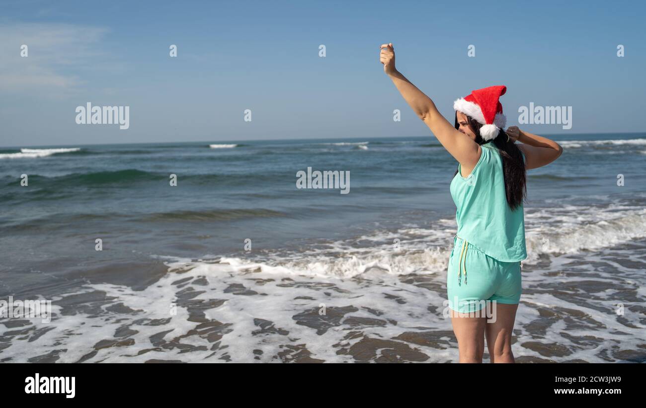 Giovane donna nel cappello di Babbo Natale in piedi in riva al mare in tempo di sole. Brunette per adulti nel cappello di Babbo Natale che si posa vicino al mare ondulato sul resort Foto Stock
