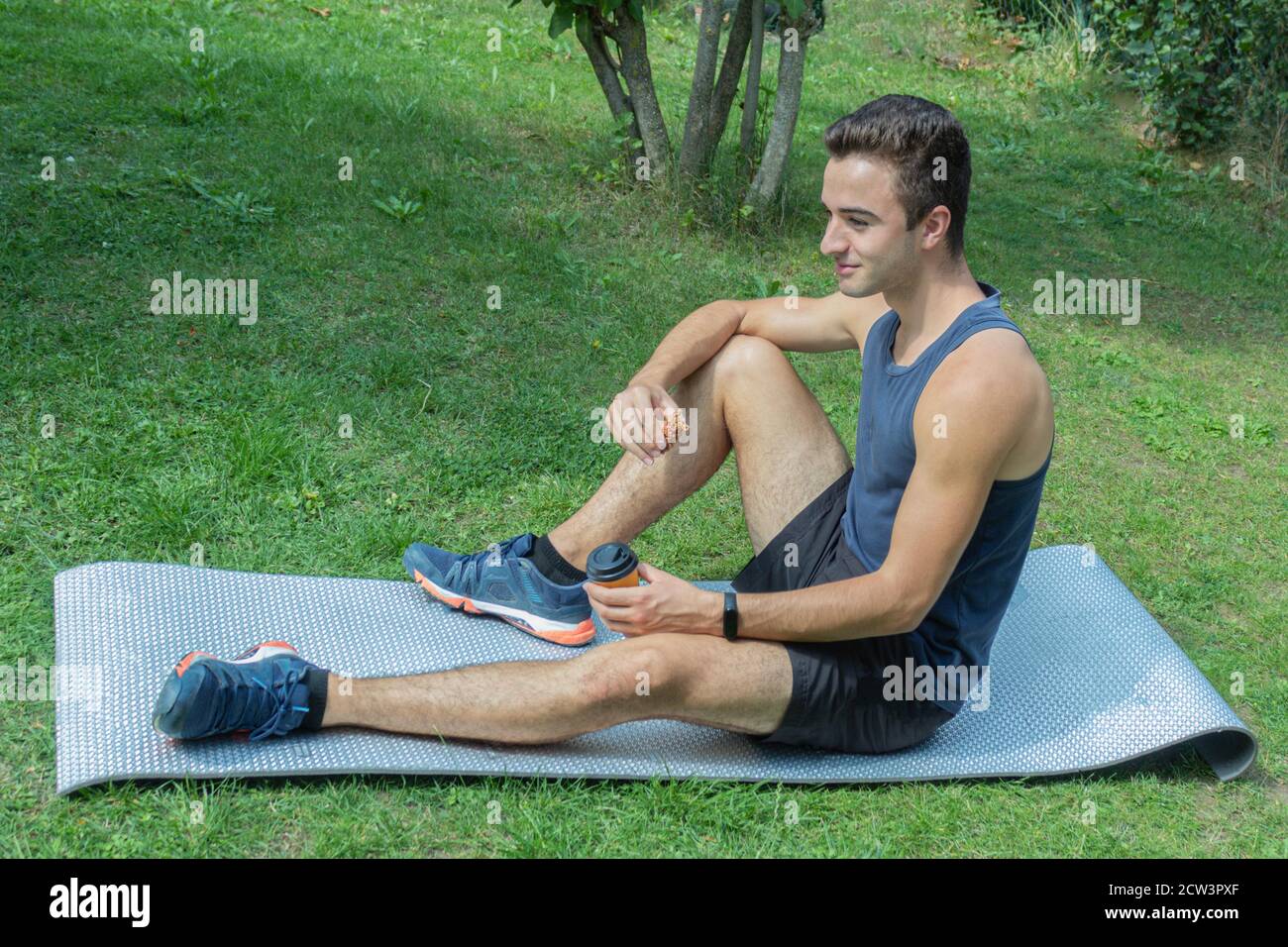 giovane atleta di fitness sorridente e seduto sul tappetino nel giardino della sua casa bevendo un piccolo caffè e un energy bar in una giornata di sole Foto Stock