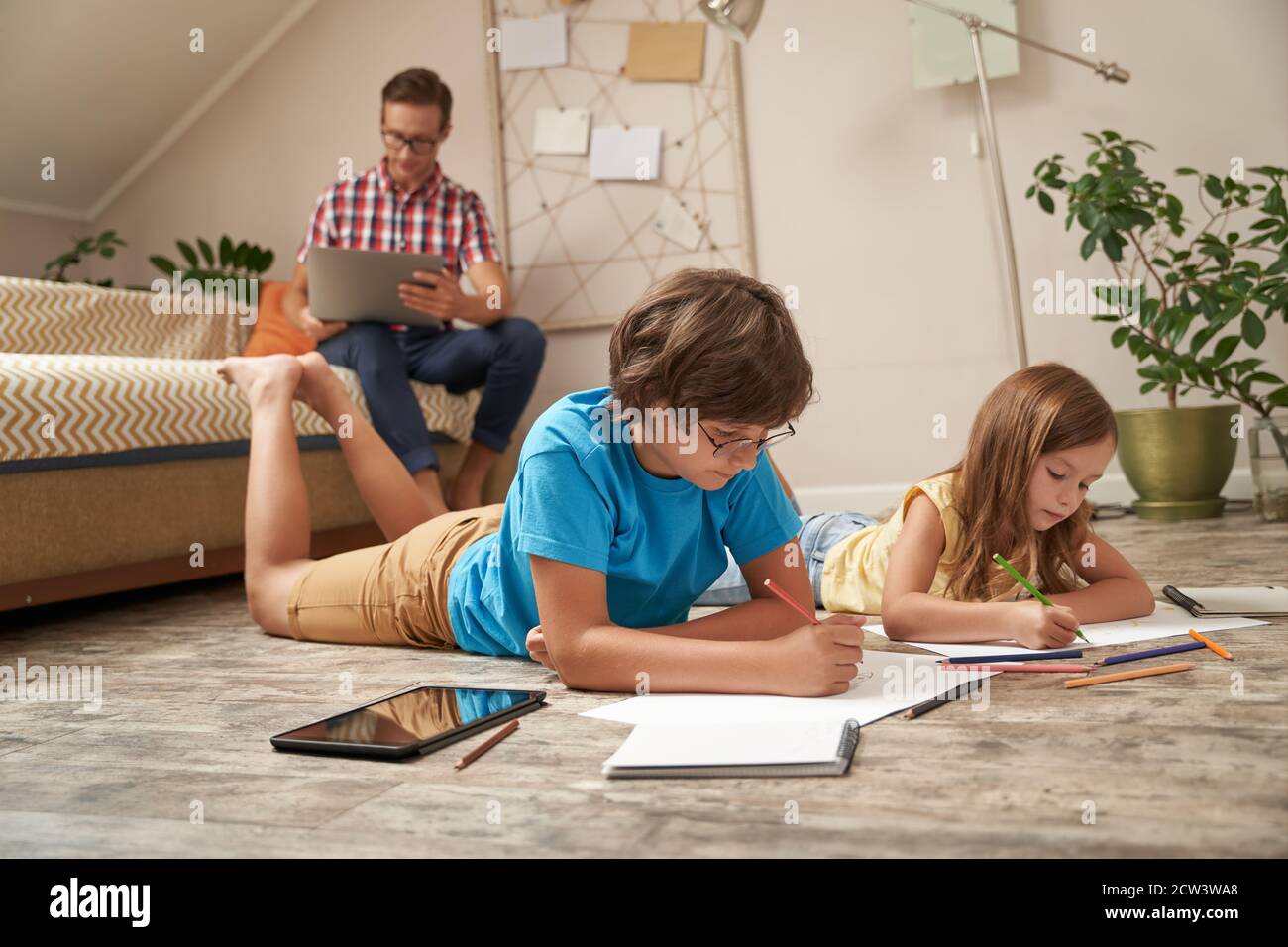 Carino ragazzino e ragazza giacente sul pavimento di legno e disegnando con matite colorate mentre il loro padre sedeva sopra divano e lavoro su computer portatile Foto Stock