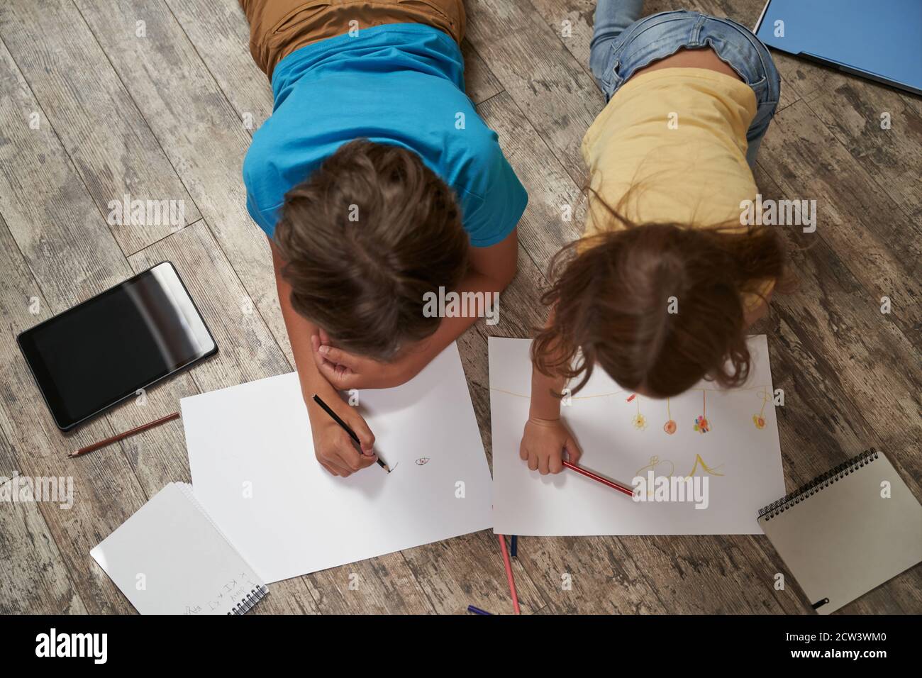 Fratelli amicizia. Vista dall'alto del piccolo fratello caucasico e della sorella giacenti sul pavimento di legno a casa e disegnati insieme su fogli bianchi di carta Foto Stock