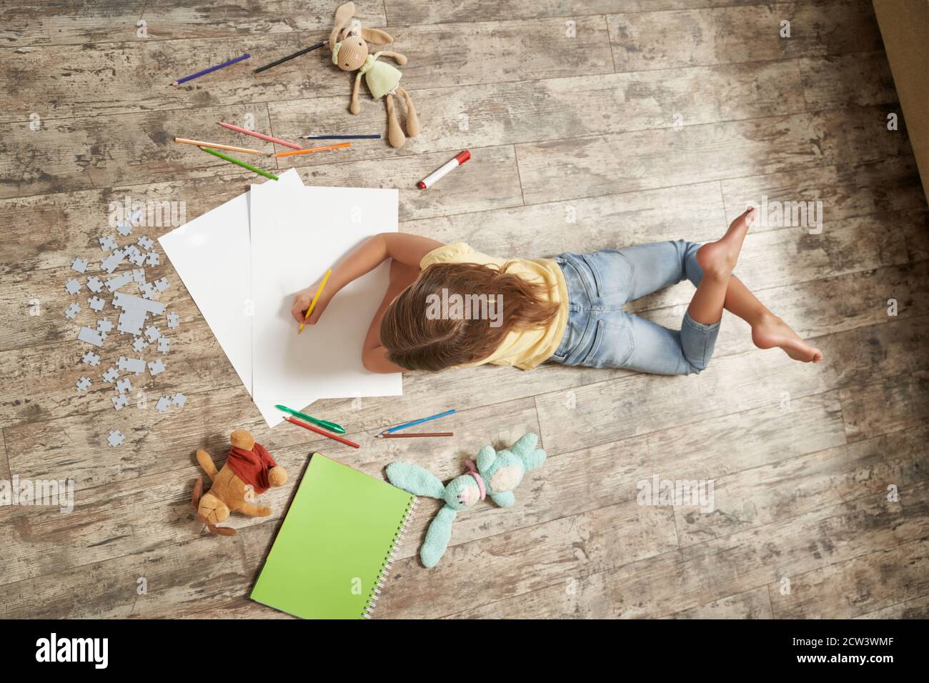 Vista dall'alto della bambina sdraiata sul pavimento in legno nella sua stanza a casa e disegnando con matite colorate su un foglio di carta bianco Foto Stock