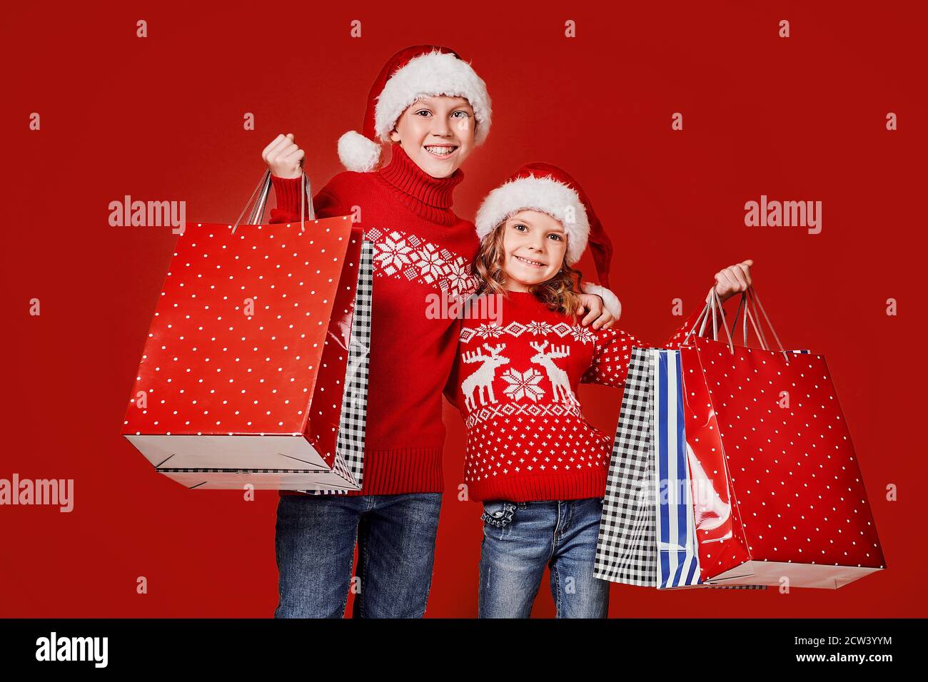 Cute bambini in rosso Santa cappelli, maglioni che tengono borse shopping su sfondo rosso. Foto Stock
