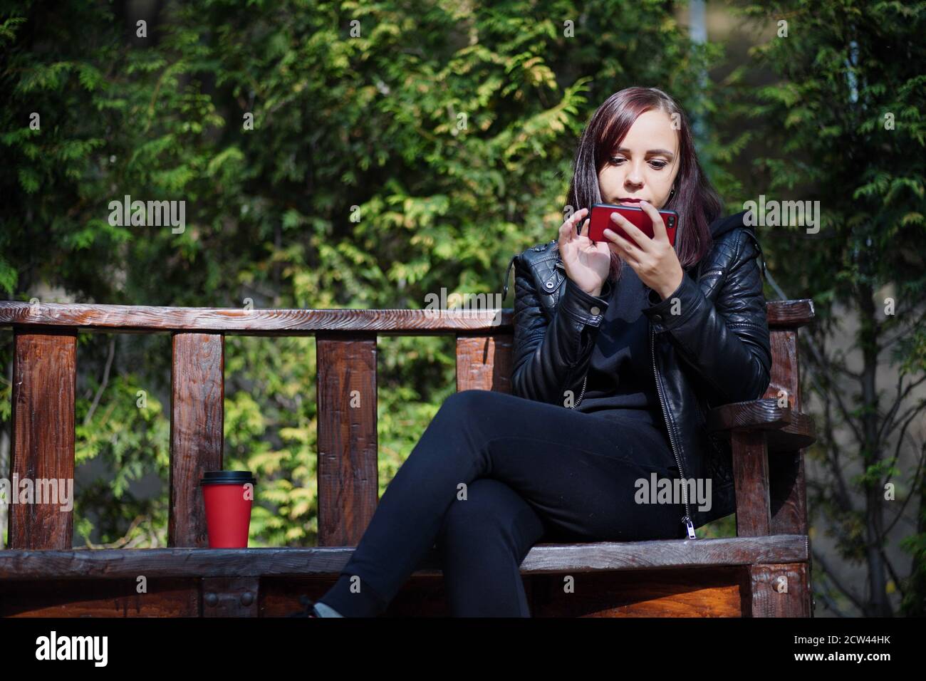 Giovane donna in giacca di pelle seduta sulla panchina e giocando sullo smartphone nel parco. La brunetta per adulti riposa tra alberi verdi nel parco cittadino e nella navigazione Foto Stock