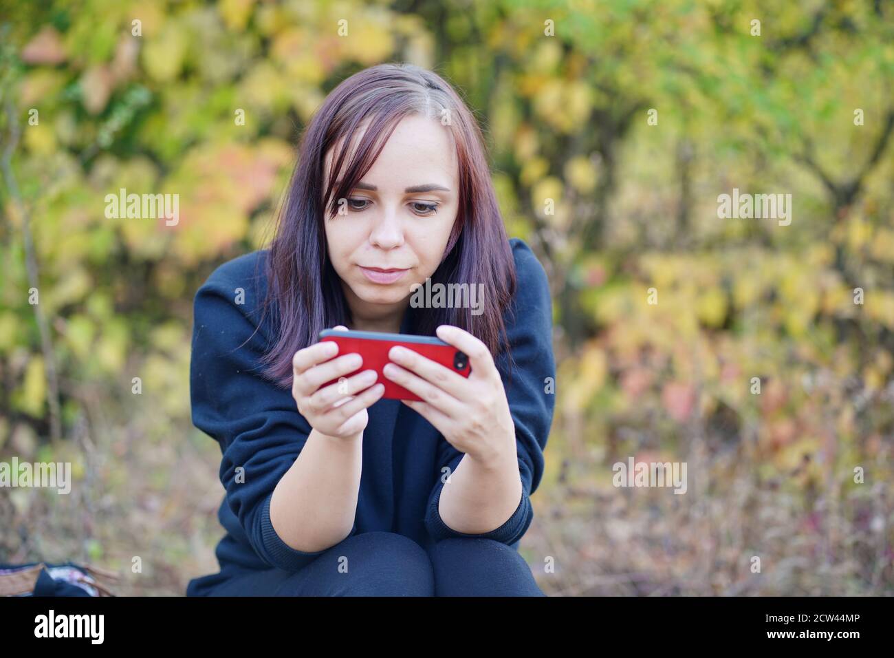 Giovane donna in abiti casual seduta su log e di navigazione cellulare. La brunetta per adulti riposa con uno smartphone sulla radura nella foresta Foto Stock