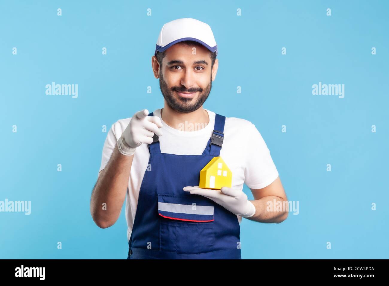 Ciao! Ritratto di felice handyman, uomo in movimento in abbigliamento da lavoro e guanti che tengono la casa di carta e che punta alla macchina fotografica. Pubblicità riparazione casa, manutenzione Foto Stock
