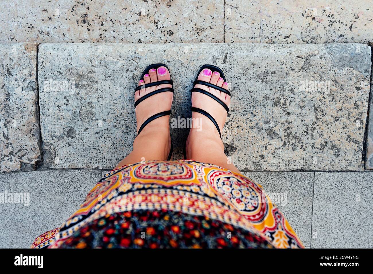 Piedi donna in prima persona vista con unghie verniciate fucsia e scarpe nere, in abito tribale marocchino, su piastrelle di pietra grigia. Concetto di etnia Foto Stock