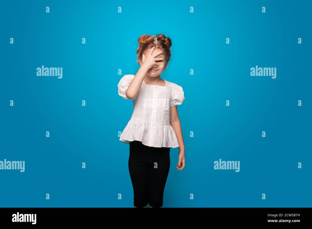 Caucasica ragazza che si nasconde usando la sua palma mentre sorride e. gesturing timido su una parete blu dello studio Foto Stock