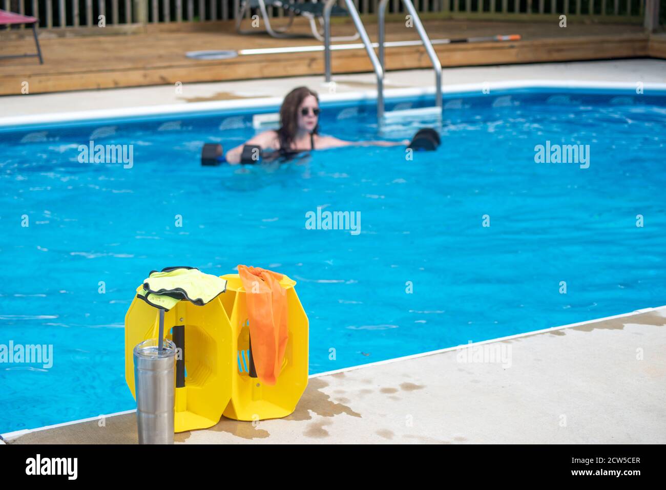 Donna che si allenava da sola in piscina con messa a fuoco sull'attrezzatura da ginnastica in acqua a bordo piscina in primo piano. Luce naturale, spazio di copia, distanza sociale Foto Stock