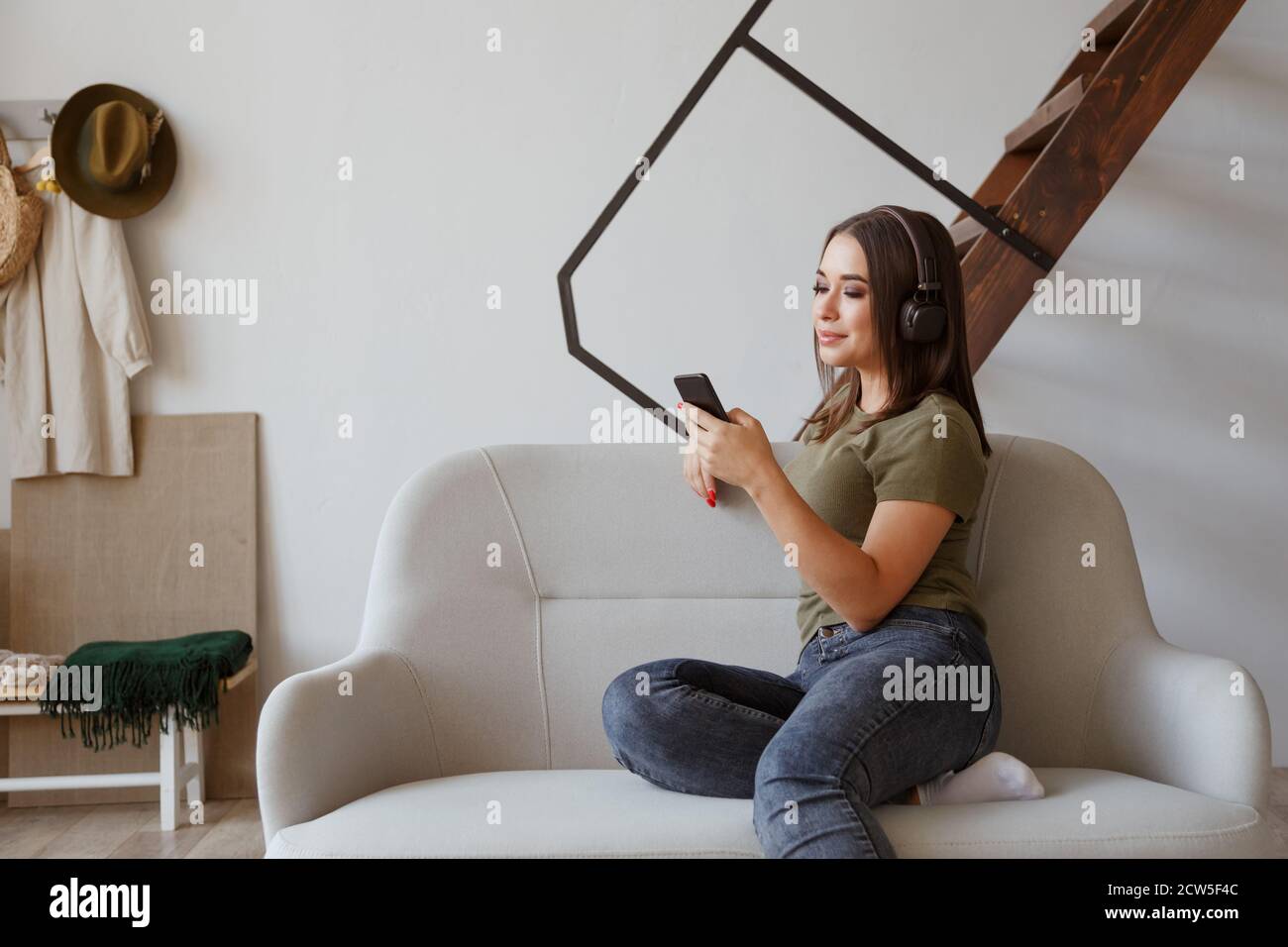 Giovane donna allegra che ascolta la musica con le cuffie wireless collegate smartphone in appartamento mansardato Foto Stock