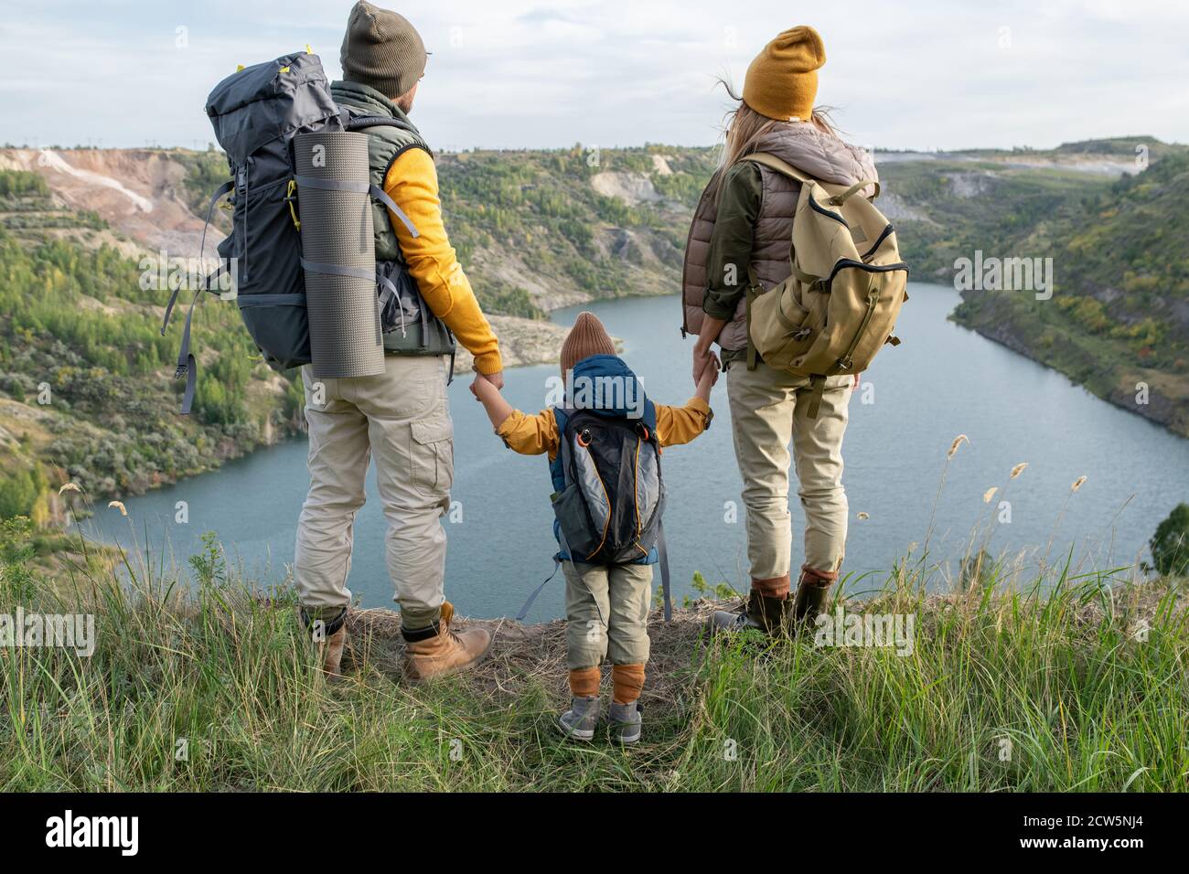 Vista posteriore dei giovani genitori e del loro figlio con zaini guardando il lago o il fiume Foto Stock