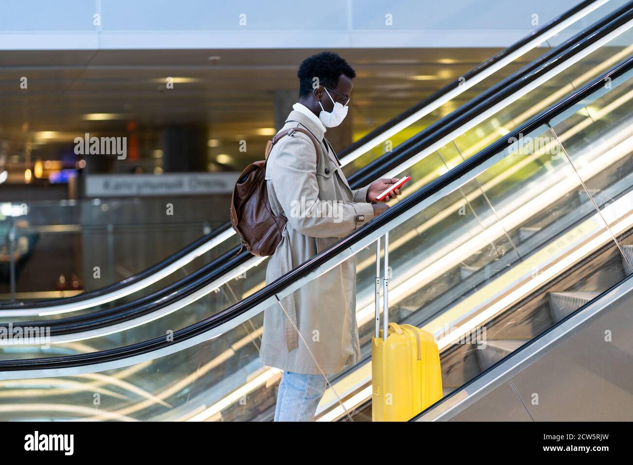 Uomo viaggiatore afro-americano con valigia gialla si trova sulla scala mobile nel terminal dell'aeroporto, indossare maschera medica viso per proteggersi dal contatto con Foto Stock