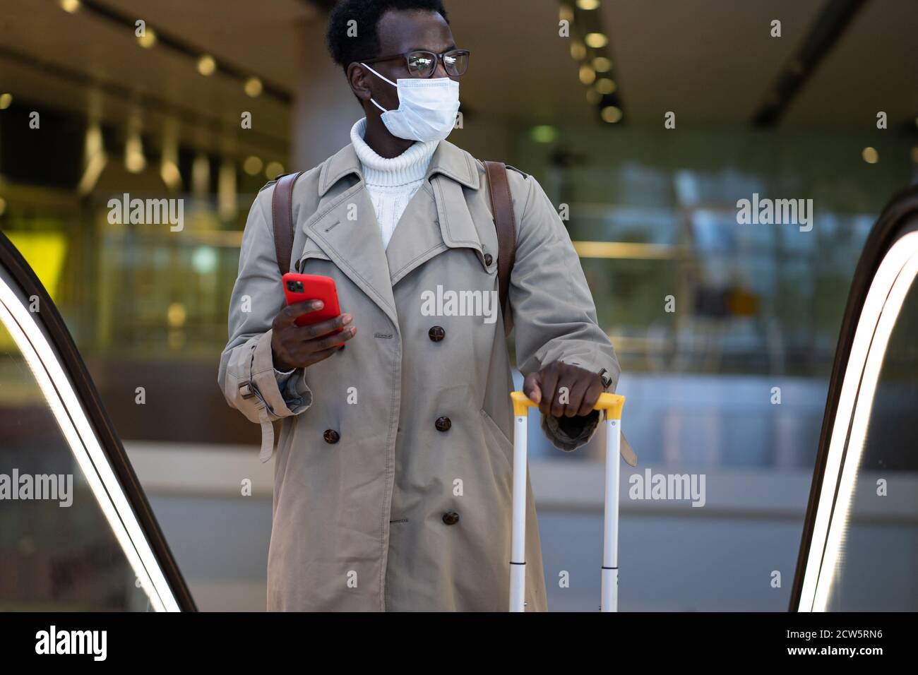Uomo viaggiatore afro-americano con valigia gialla si trova sulla scala mobile nel terminal dell'aeroporto, indossare maschera medica viso per proteggersi dal contatto con Foto Stock