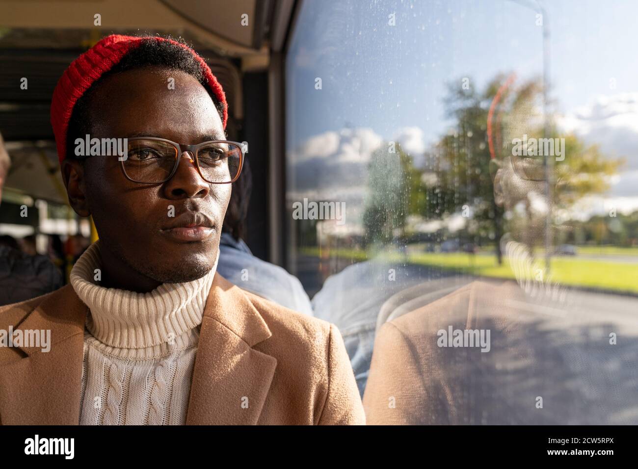 Ritratto dell'uomo viaggiatore afro-americano messo a fuoco in camice beige, maglione bianco e cappello rosso, indossando occhiali, guardando la finestra, viaggiando dal pubblico Foto Stock