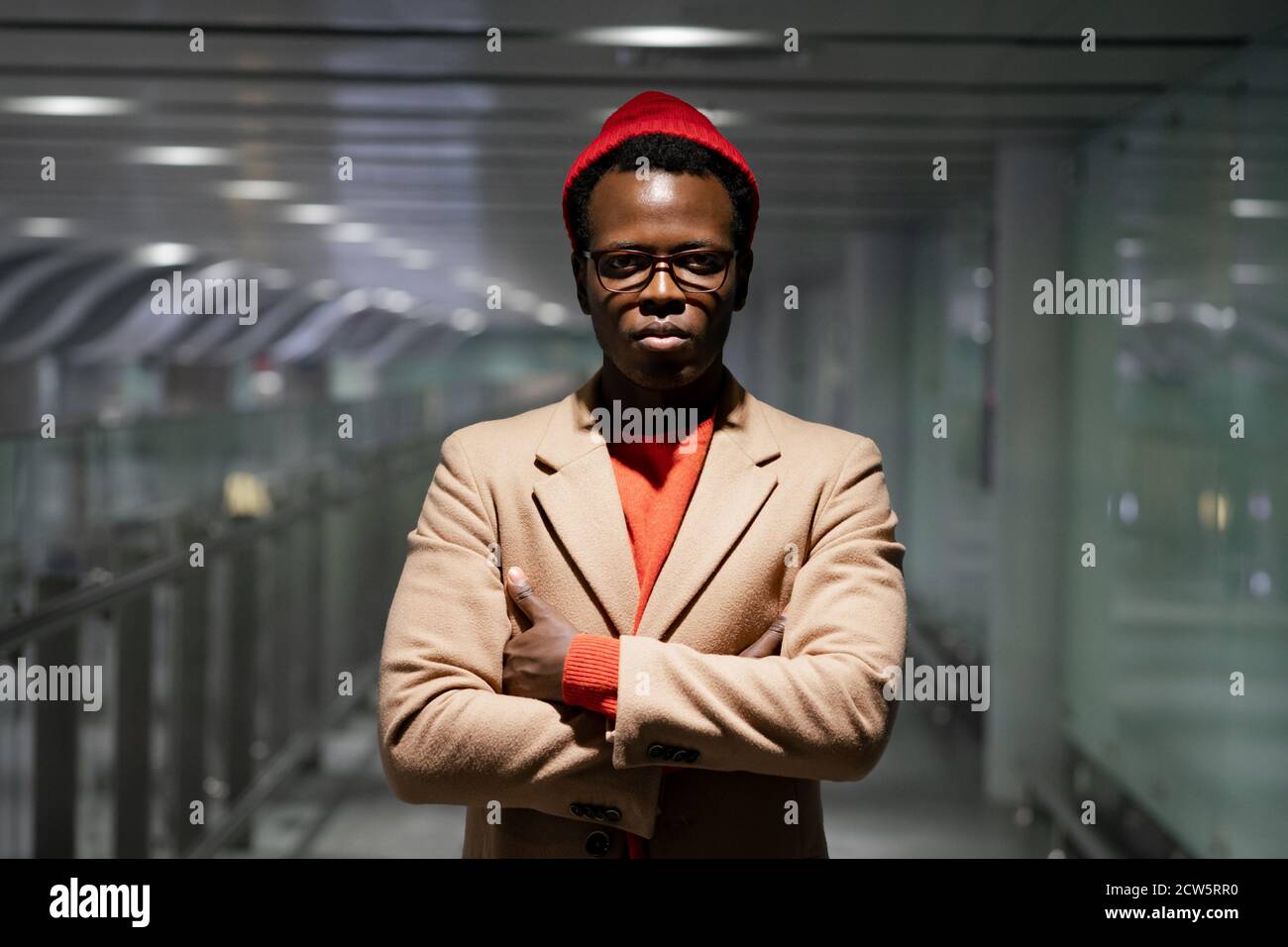 Elegante uomo millenario afro-americano in occhiali indossare cappotto beige, maglione rosso e cappello, in piedi nella sala dell'aeroporto, guardando la fotocamera, le braccia incrociate Foto Stock
