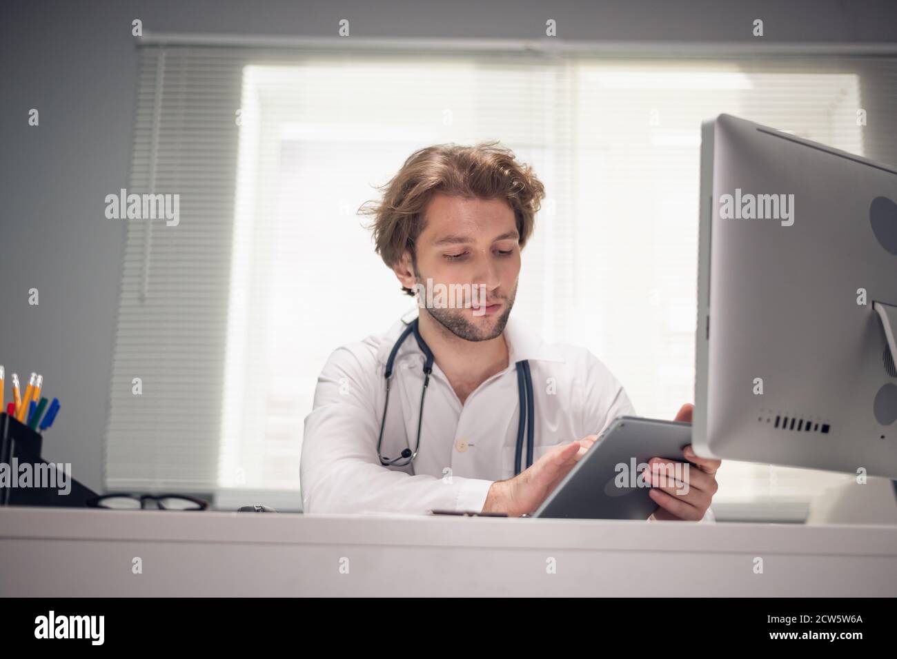 Un ritratto di un medico seduto con un tablet digitale alla sua scrivania, controllando i messaggi, facendo correzioni nel programma. Foto Stock