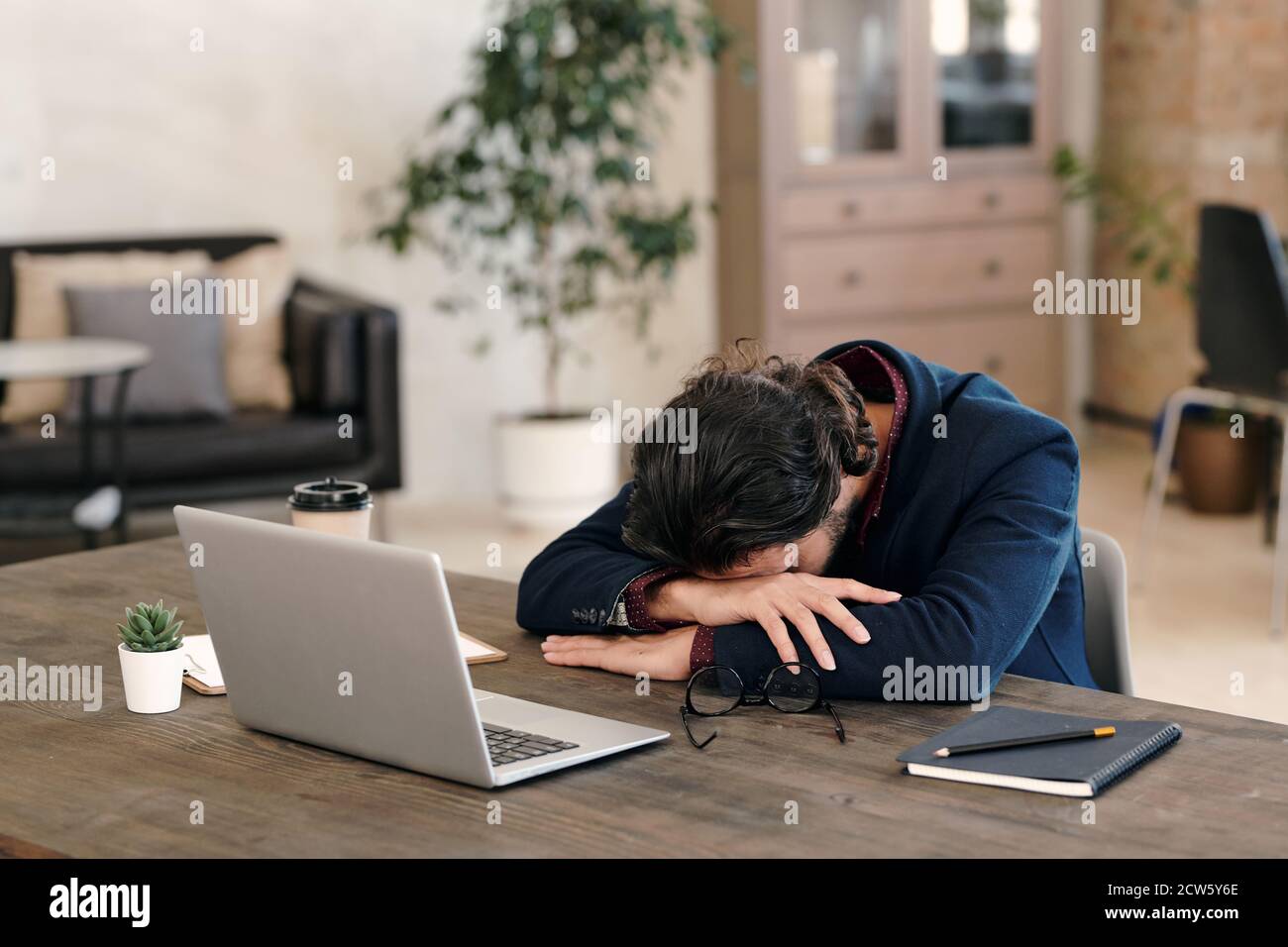 Giovane uomo d'affari stanco in formalwear che tiene la testa sulle braccia durante il napping Foto Stock