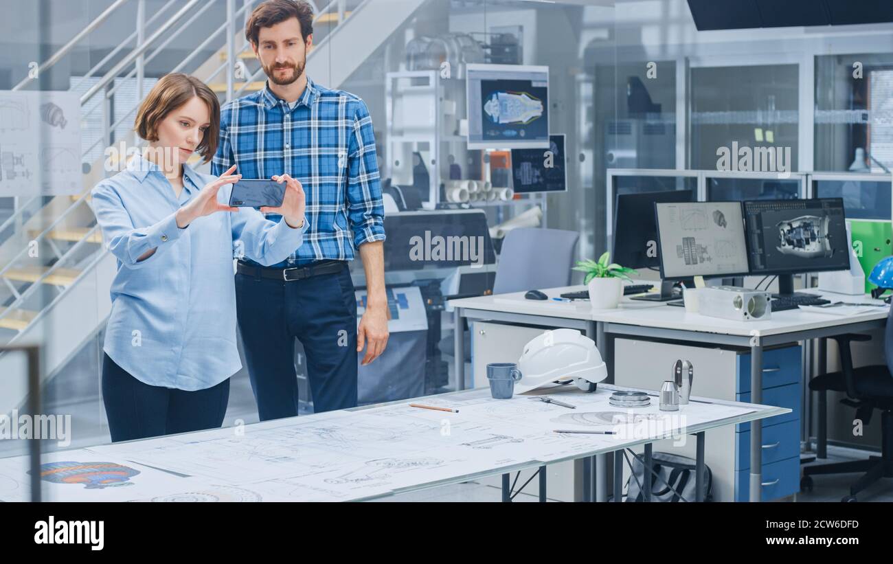 Nello stabilimento di ingegneria industriale: La designer femminile con capo ingegnere utilizza lo smartphone per l'app di realtà aumentata per manipolare la progettazione di motori Foto Stock