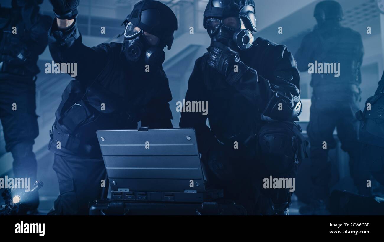 Il team mascherato degli ufficiali di polizia SWAT armati con i fucili si trovano in Dark Gipped Office Building con scrivanie e computer. Soldier apre un computer portatile a. Foto Stock