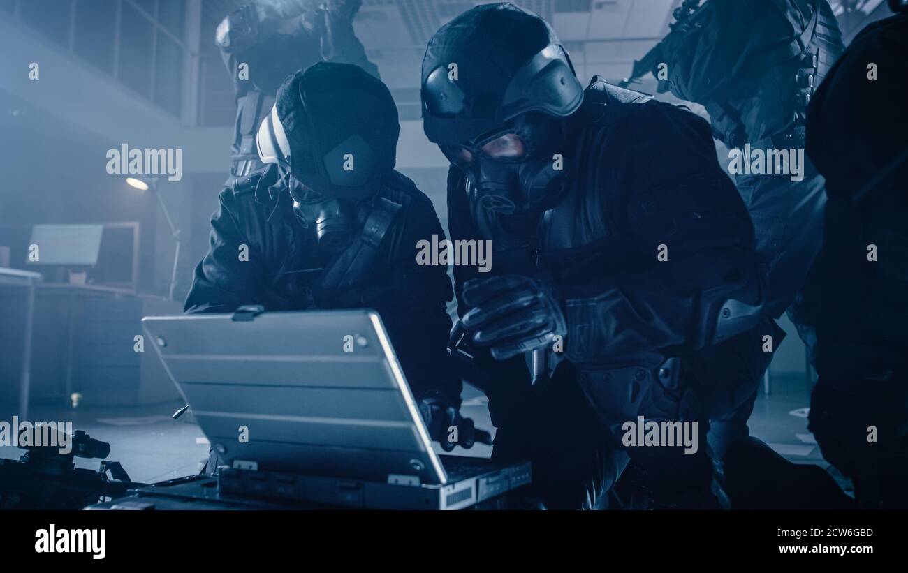 Il team mascherato degli ufficiali di polizia SWAT armati con i fucili si trovano in Dark Gipped Office Building con scrivanie e computer. Soldier apre un computer portatile a. Foto Stock