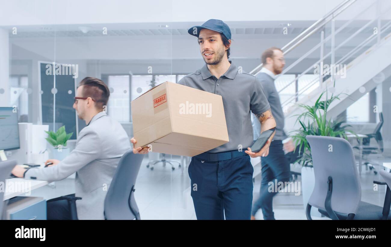 Happy Delivery Man entra nell'ufficio aziendale e consegna il cartone a un lavoratore. Grande luminoso moderno ufficio aziendale con professionista Foto Stock