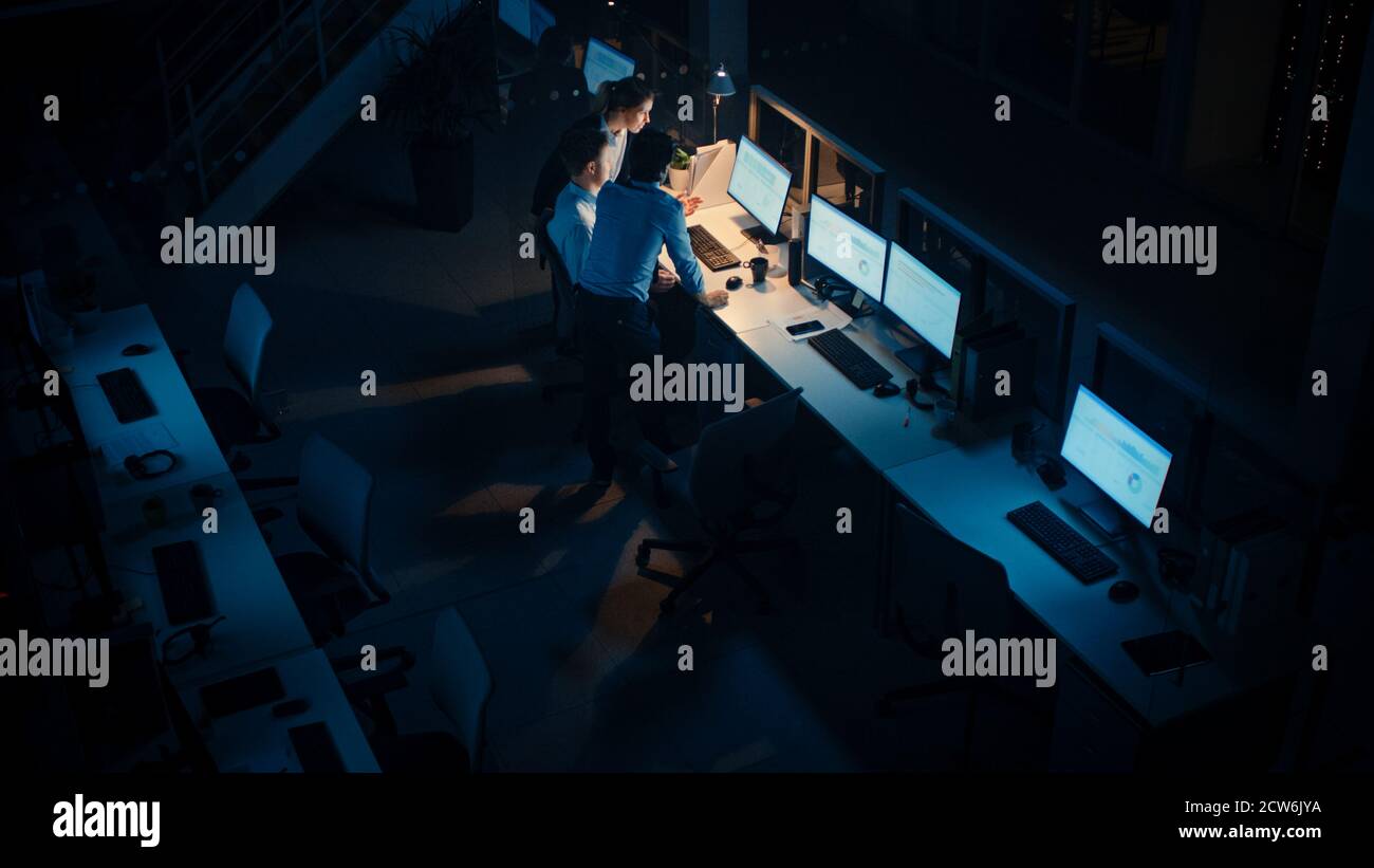 Tarda notte in ufficio moderno: Team diversificato di uomini d'affari di successo e donne d'affari si riuniscono intorno al computer, hanno discussione, trovare problema Foto Stock