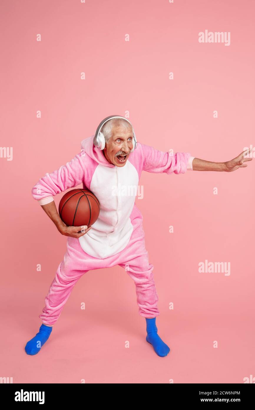 Giocare a basket con le cuffie. Ritratto di uomo hipster senior in elegante  abbigliamento isolato su sfondo rosa studio. Concetto di stile di vita per  anziani con tecnologia e gioia. Colori alla