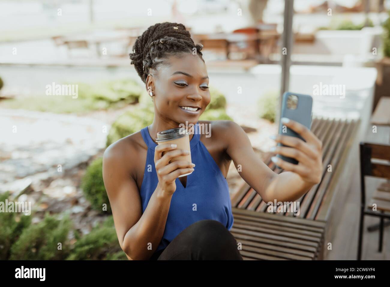 Allegra donna africana passeggiate all'aperto, tenendo takeaway tazza da caffè, prendendo un selfie seduta sul banco di lavoro Foto Stock