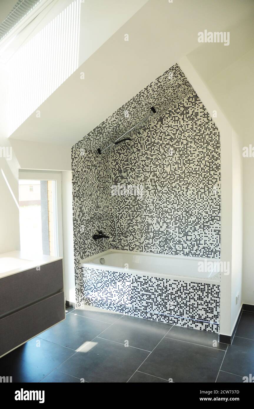 Moderno design mansarda con pareti in mosaico di vetro bianco e nero, bagno nero opaco con rubinetto e doccia, vasca bianca e una finestra. Foto Stock