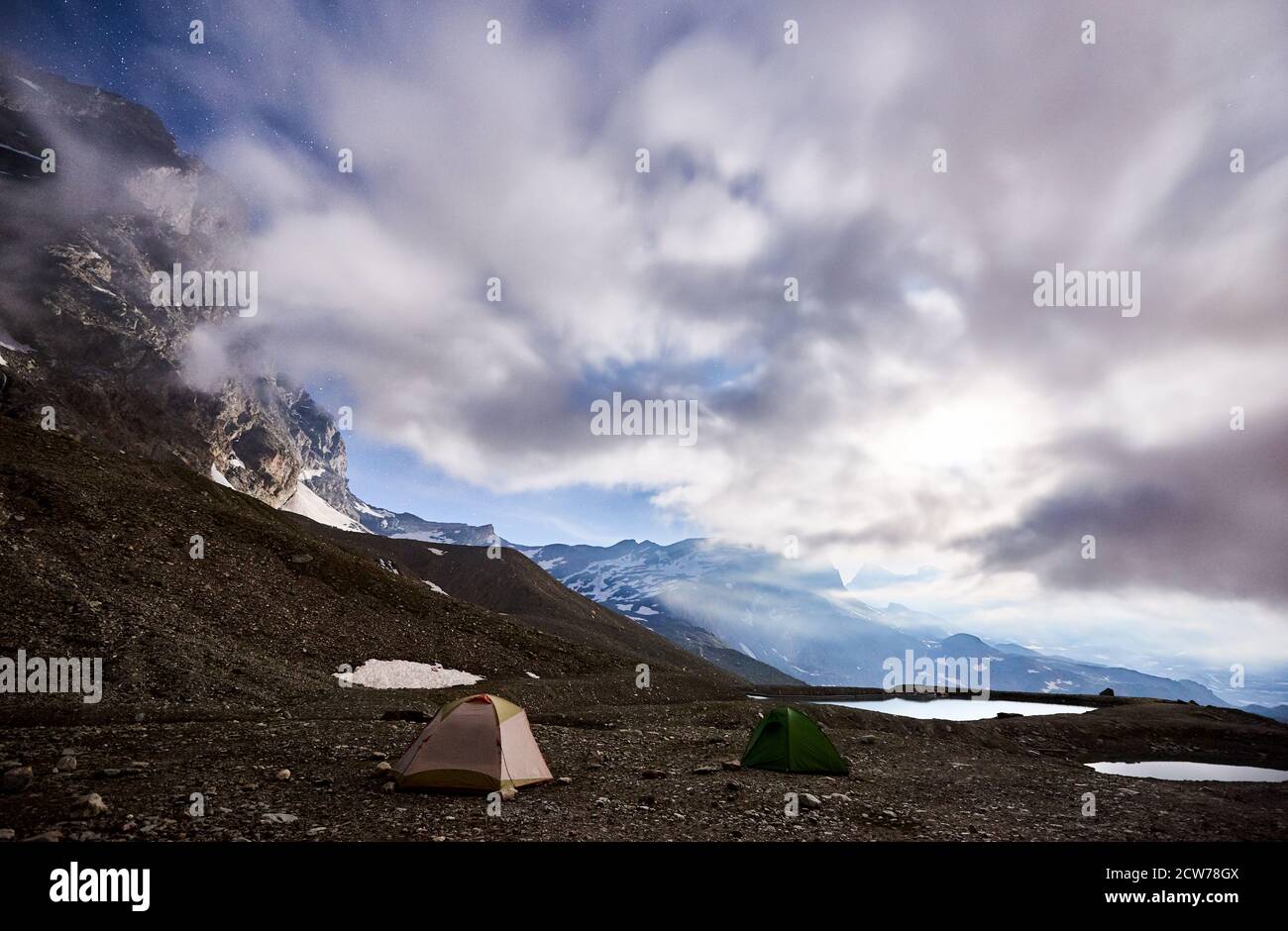 Magico scenario di cielo blu nuvoloso e stelle splendenti, bella zona di montagne con piccoli laghi. Splendida cresta di montagna con alte vette rocciose, due colorate tende turistiche. Campeggio notturno nelle Alpi Foto Stock