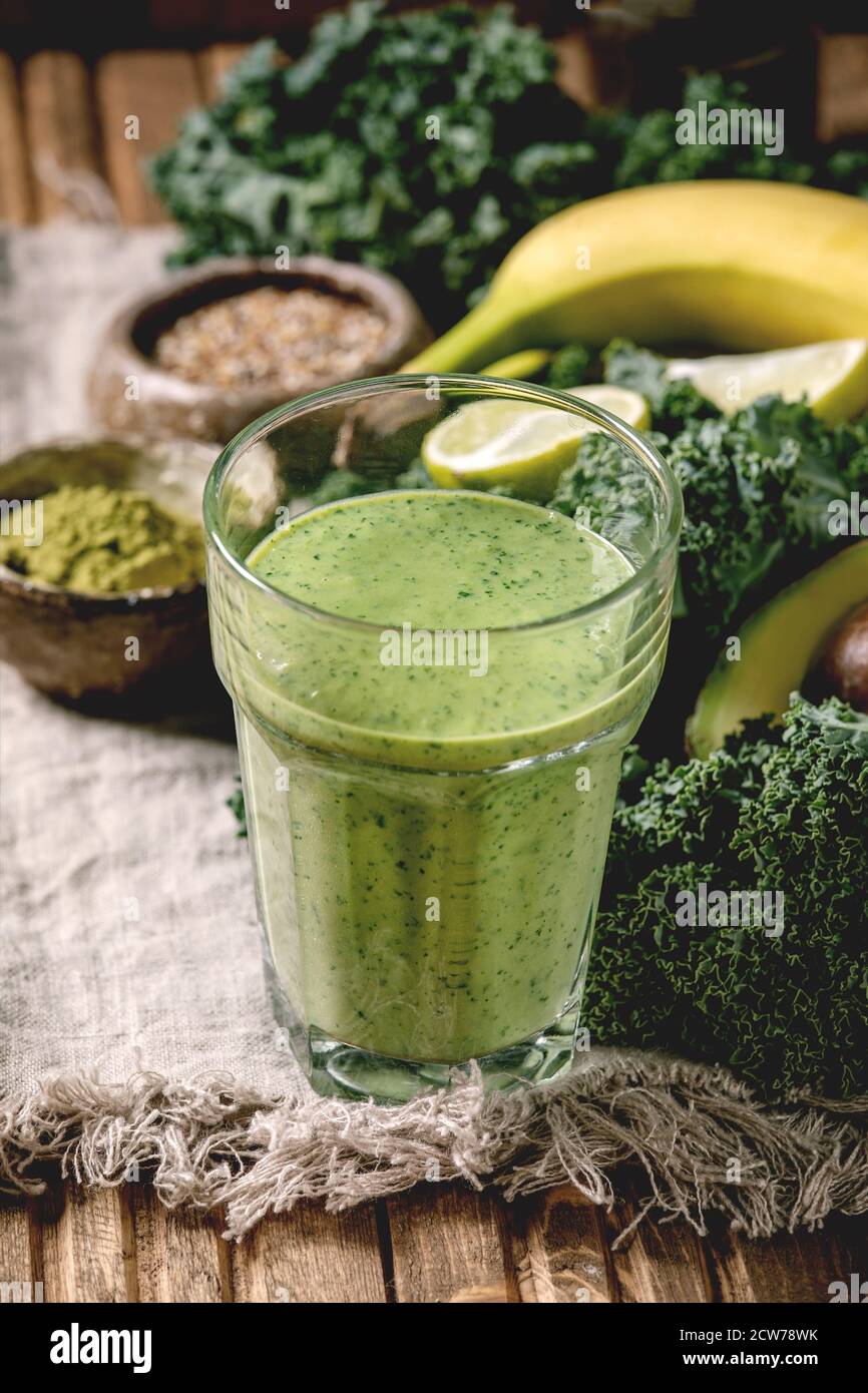 Bicchiere di frullato vegano verde sano, paglia di vetro. Ingredienti sopra. Kale, banane, avocado, calce, latte non-diario, polvere di matcha e semi sul legno Foto Stock