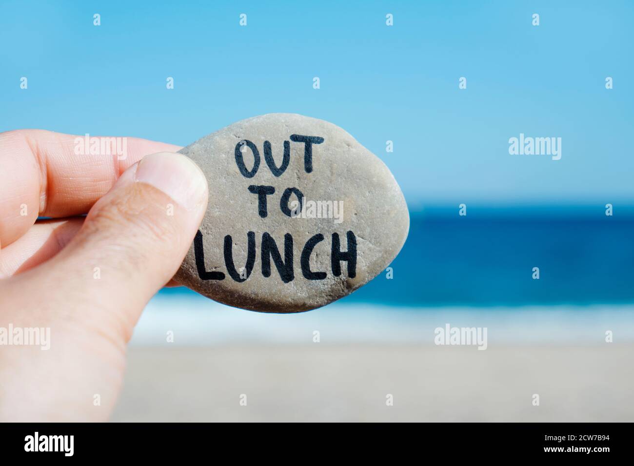 primo piano della mano di un giovane caucasico sulla spiaggia, di fronte all'oceano, tenendo una pietra con il testo scritto a pranzo Foto Stock