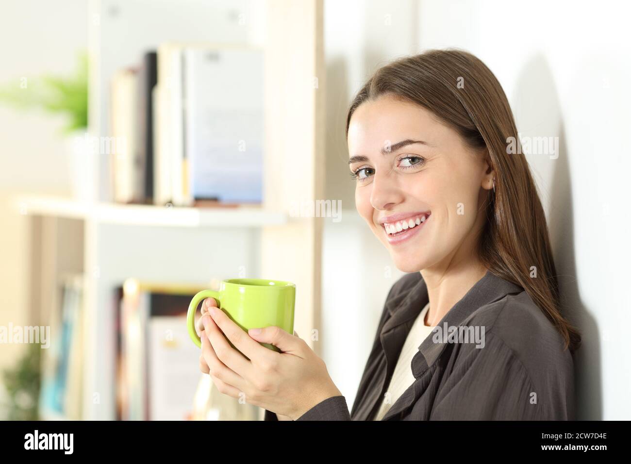 Donna felice che tiene la tazza di caffè verde guarda la macchina fotografica in piedi a casa Foto Stock