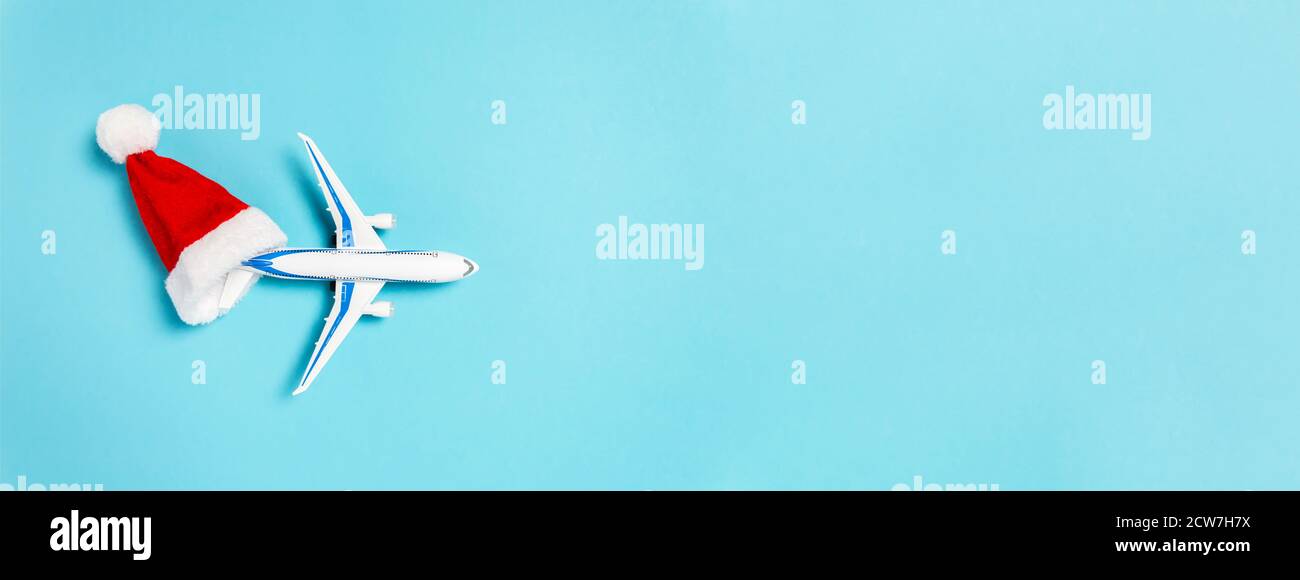 Banner natalizio con concetto di viaggio per le vacanze. Aereo bianco che vola dal cappello rosso di Babbo Natale su sfondo blu. Stile piatto compositio minimo Foto Stock