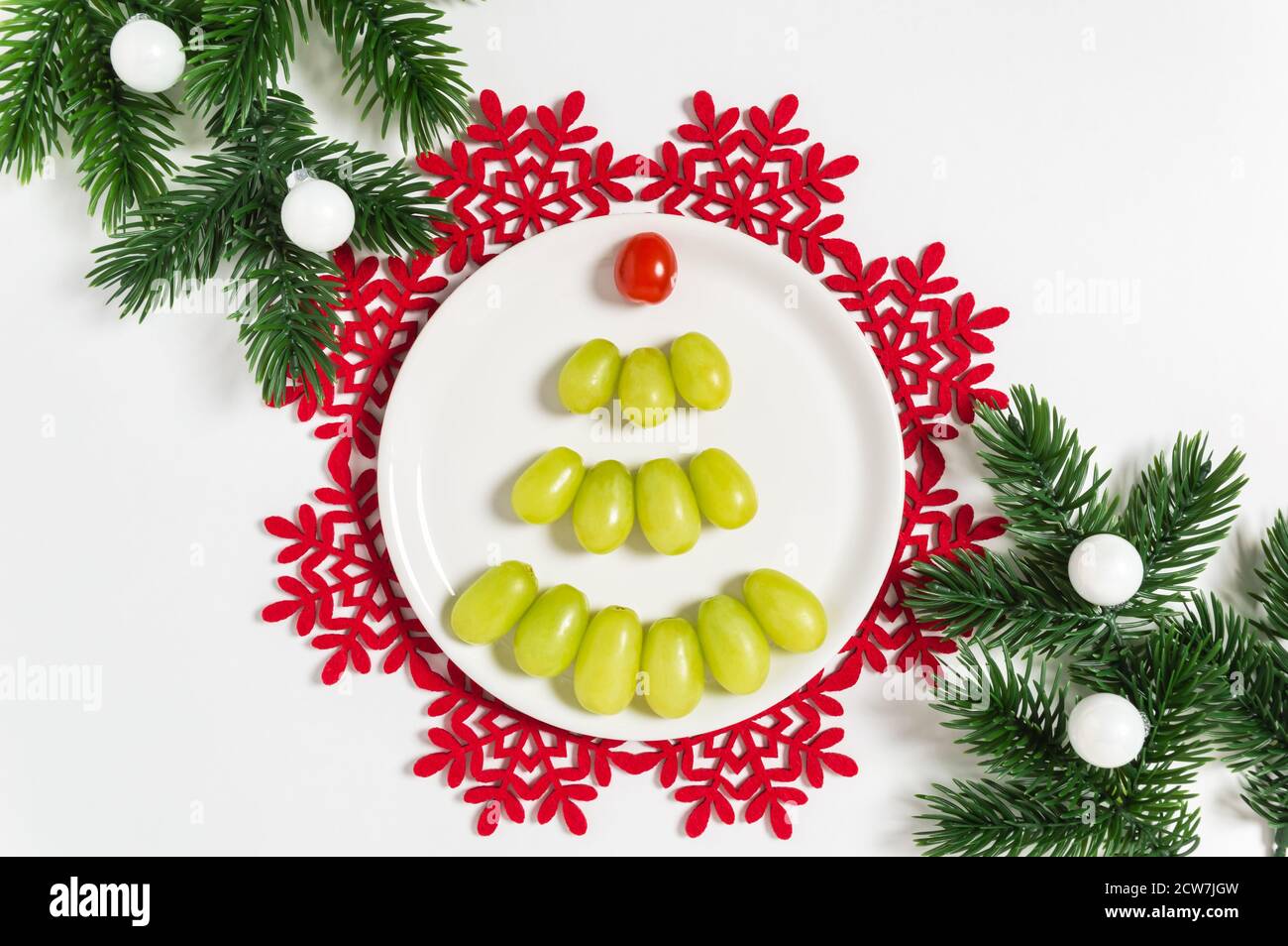 Albero di Natale di uve verdi e pomodori ciliegini con rami di pino e palle ornamentali. Frutta sul tavolo di Natale. Fondo alimentare di Capodanno t Foto Stock