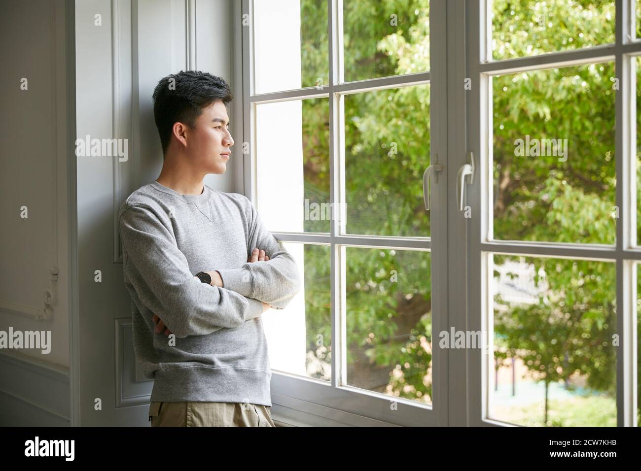 giovane uomo asiatico in piedi dalla finestra a casa braccia incrociate Foto Stock