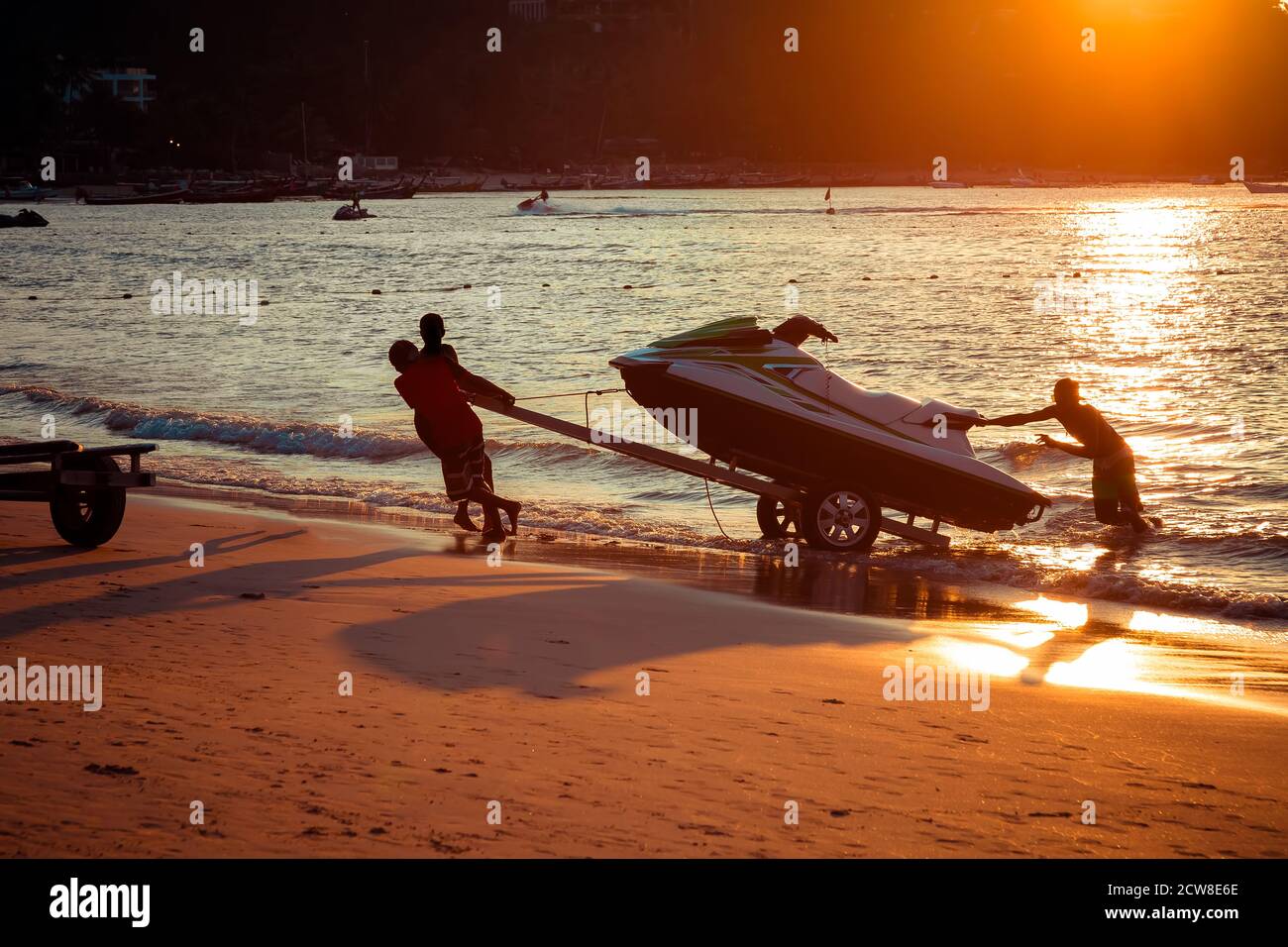 Gli uomini al tramonto tirano l'idroticlo fuori dall'acqua su una spiaggia di sabbia. Vacanze estive. Moto d'acqua caricata su un rimorchio Foto Stock