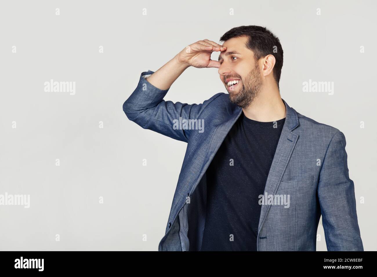 Giovane uomo d'affari con barba in giacca, molto felice e sorridente, guardando lontano con la mano sopra la testa. Concetto di ricerca. Ritratto di un uomo su sfondo grigio. Foto Stock