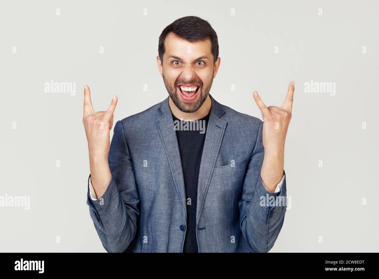 Giovane uomo d'affari con barba in giacca, gridando con un'espressione pazza sul viso, facendo un simbolo rock con le mani in su. Stella della musica. Concetto di musica pesante. Uomo su sfondo grigio. Foto Stock