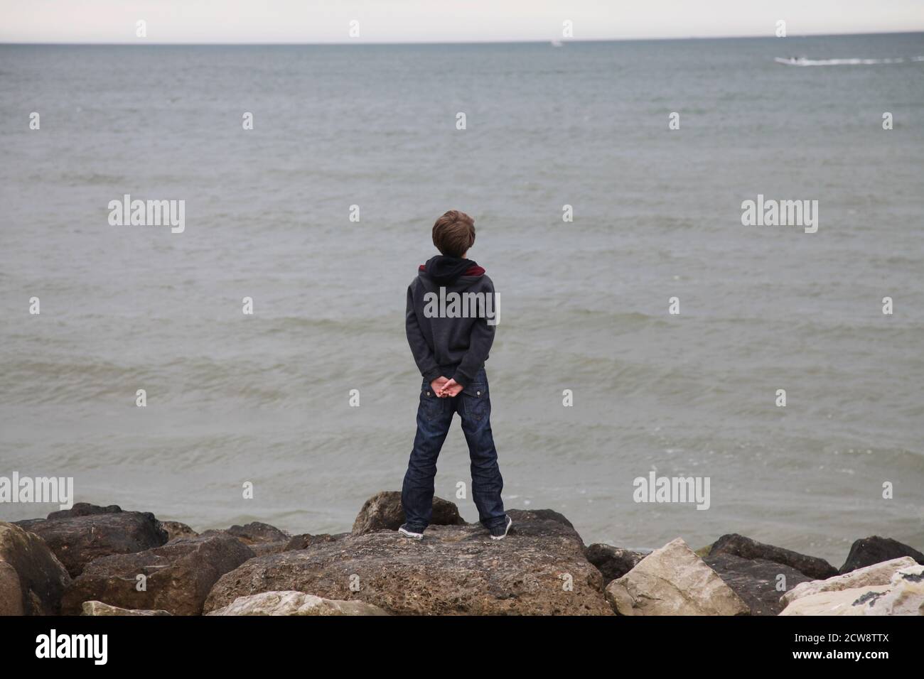 Lone boy in piedi su un groyne sulla costa sud inglese, guardando una barca verso il mare Foto Stock