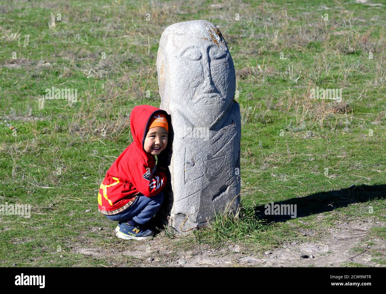 Bambino kirghiso che si nasconde dietro un antico bal-bal, una lapide usata dalle tribù nomadi turche vicino a Balasagun in Kirghizistan, Asia centrale. Ragazzo sorridente. Foto Stock