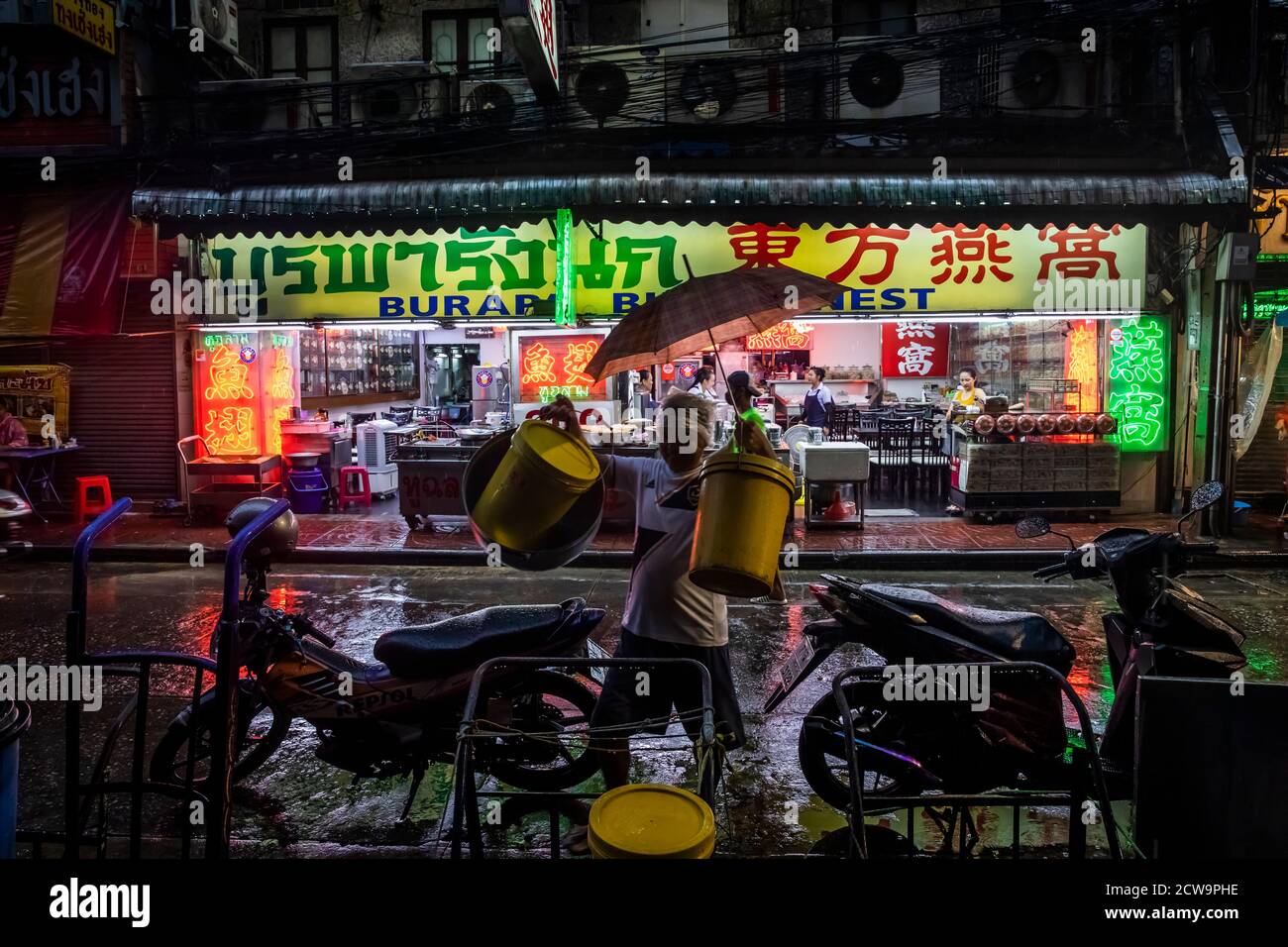 Un operaio locale che porta due secchi gialli attraversa la strada di fronte a un mercato nella Chinatown di Bangkok. Foto Stock