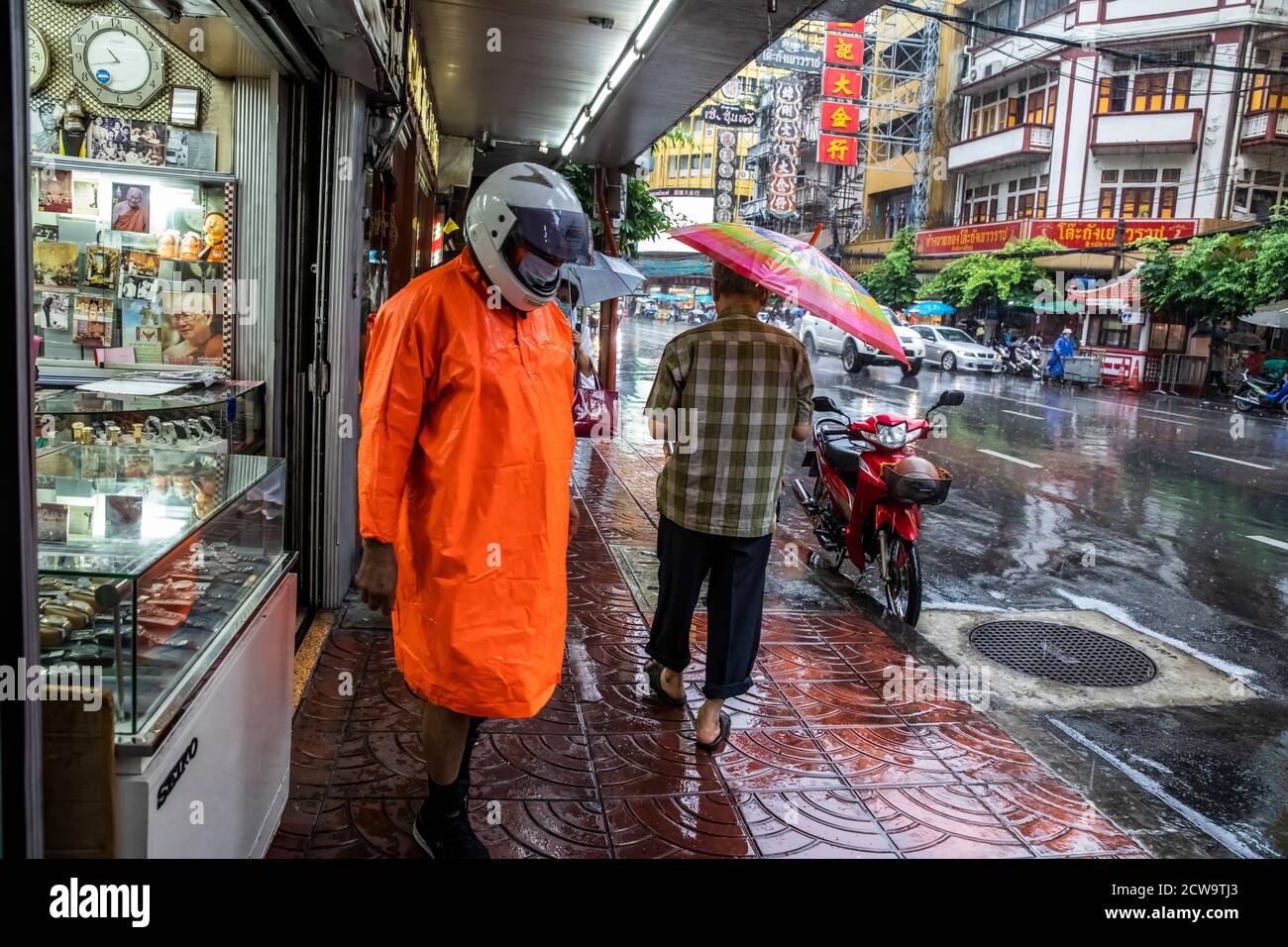 Un uomo dawns impermeabile e helmut a cavalcare la sua moto sotto la pioggia mentre un altro passa con un ombrello nella Chinatown di Bangkok. Foto Stock