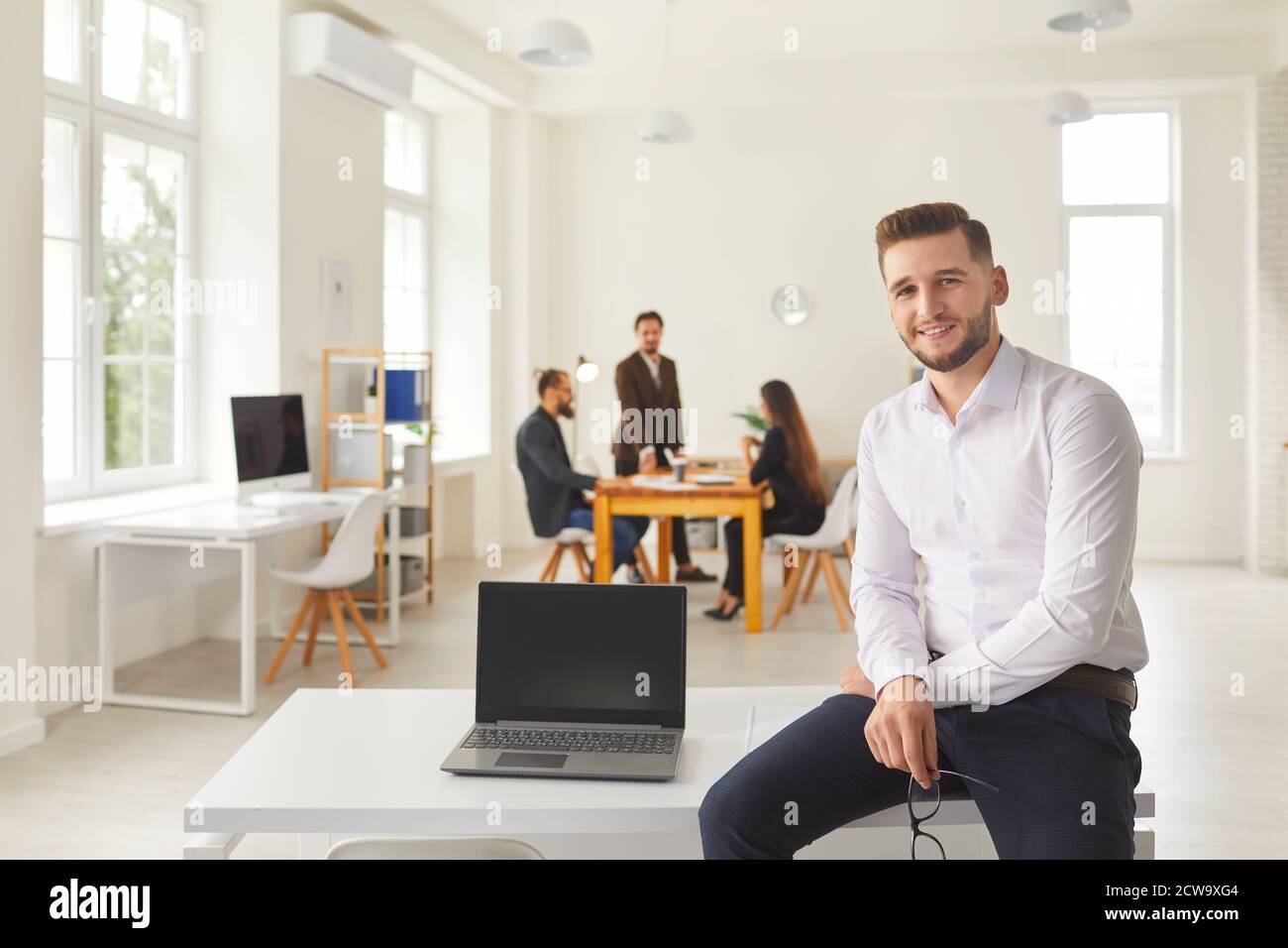 Giovane uomo d'affari con computer portatile con schermo vuoto seduto sulla scrivania nello spazio dei colleghi guardando la fotocamera Foto Stock