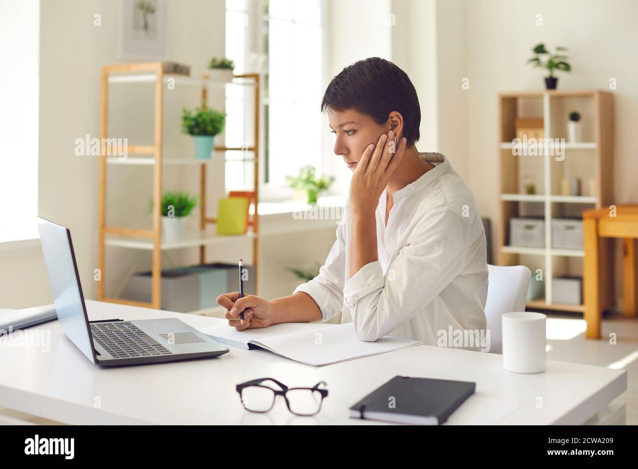 Donna concentrata con notebook e penna che lavora in un elegante e luminoso ufficio su un computer portatile. Foto Stock